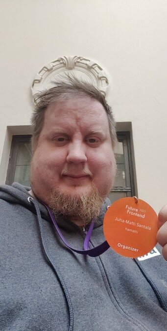 Me wearing a dark grey hoodie and holding a bright orange organizer badge for Future Frontend conference with my name on it.