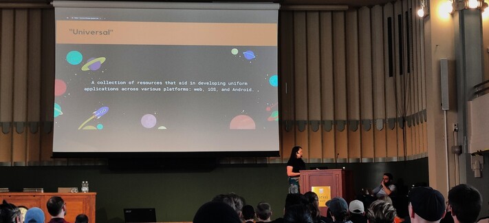 Thais on stage with a slide describing universal design system: "A collection of resources that aid in developing uniform application across various platforms: web, iOS and Android"
