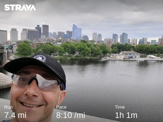 selfie with my glasses and Boston Marathon hat, with Boston skyline and the Charles River in the background