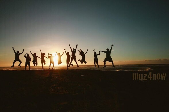 A line of people are silhouetted by the setting sun. The scene is a beach at the water's edge and each person is jumping, dancing, and waving their arms in the air. The image celebrates community.