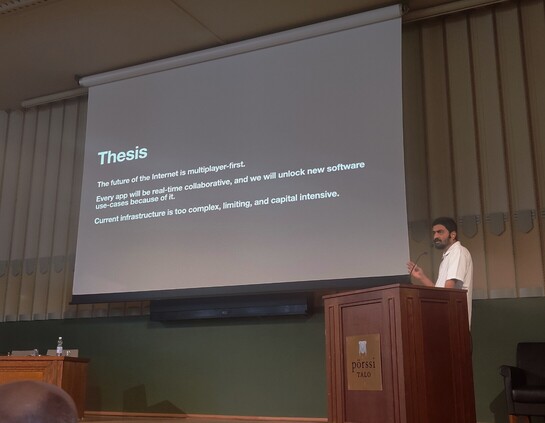 Sunil standing behind a speaker podium speaking, with a slide where he introduces his thesis of future of the internet being multiplayer
