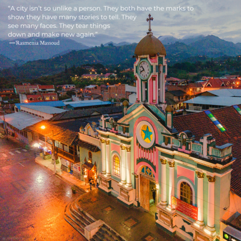 A colorful church features prominently in the photo, among small buildings alongside it in the city street. Houses and other buildings are seen in the distance at dusk, with lush green mountains and some low clouds in the background. The church is made up of the vibrant colors of seafoam green, pink, and white with some yellow accents in the stained glass windows. The quote on the photo reads, “A city isn’t so unlike a person. They both have the marks to show they have many stories to tell. They see many faces. They tear things down and make new again.” by Rasmenia Massoud from Broken Abroad.