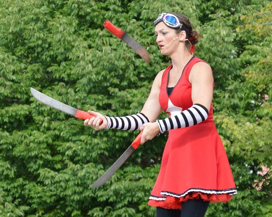 A photo of a woman juggling three knives. She is wearing a red dress, black and white striped arm warmers, and a pair of googles pushed up on her head. The background of the photo is entirely filled with green leaves of trees.