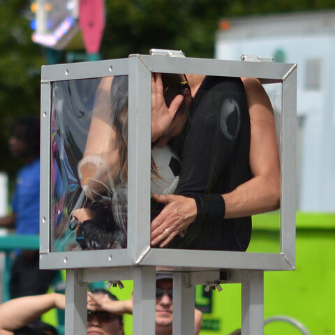 A photo of a woman contorted into a small clear box raised up on a metal stand. There is also a soccer ball in the box with her.