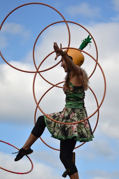 A photo of a woman in a green floral dress standing on a support with one leg. She is wearing a yellow helmet with artificial green leaves coming out of the top and spinning five hula hoops . She is spinning two in each hand and one with the leg she isn't standing on.