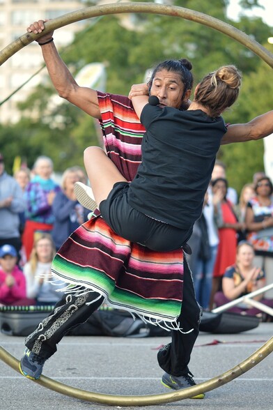 A photo of a woman with her arms and legs wrapped around a man wearing a colorful poncho. The man is spinning them in a cyr wheel.