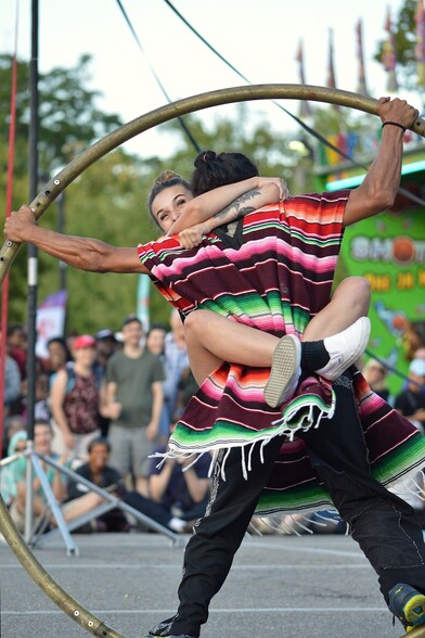 A photo of a woman with her arms and legs wrapped around a man wearing a colorful poncho. The man is spinning them in a cyr wheel.