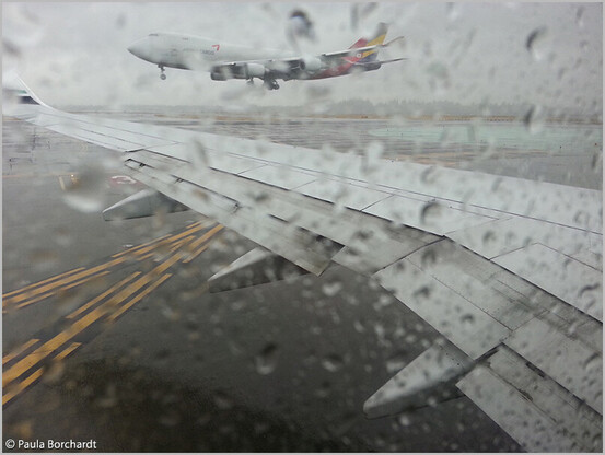 My photograph of a rainy Seattle airport runway with a plane mid-air and the wing of the plane I'm on, as seen through my rain-streaked plane window.