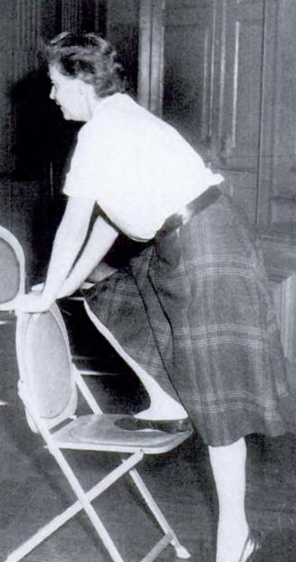 Betty Meredith-Jones teaching a movement class in the 1950s; a white woman wearing a light blouse and a plaid skirt, with two hands and one foot resting on a folding chair