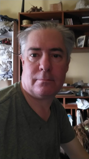 Selfie of a white guy with slicked-back grey hair and a soft chin in a green v-neck t-shirt looking right at the camera, his face lit from the side by light from a window. It's about ten in the morning. He is neither smiling nor frowning.
