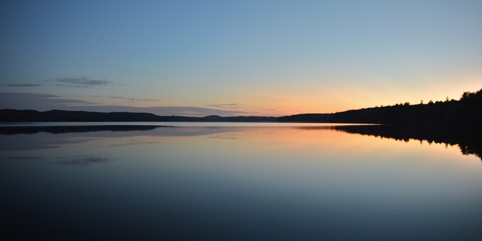 A photo of a lake at dusk. The lake is bordered by the silhouettes of low forest covered hills. The sky is mainly clear with some clouds on the horizon.