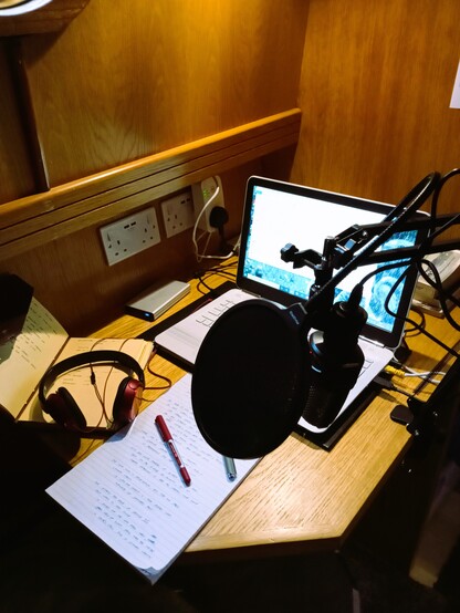 A photograph of the 'studio' I set up each week in the little study at the back (stern) of the 'Erica.' It shows a microphone, laptop, headphones, and a lot of notebooks (essential!!). 