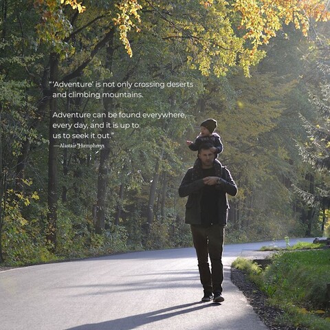 A father and son walk along a small, paved, forested road. The boy is riding up on his dad's shoulders, pointing at something in the trees off camera. The quote on the photo reads, ‘Adventure’ is not only crossing deserts and climbing mountains. Adventure can be found everywhere, every day, and it is up to us to seek it out.” by Alastair Humphreys.