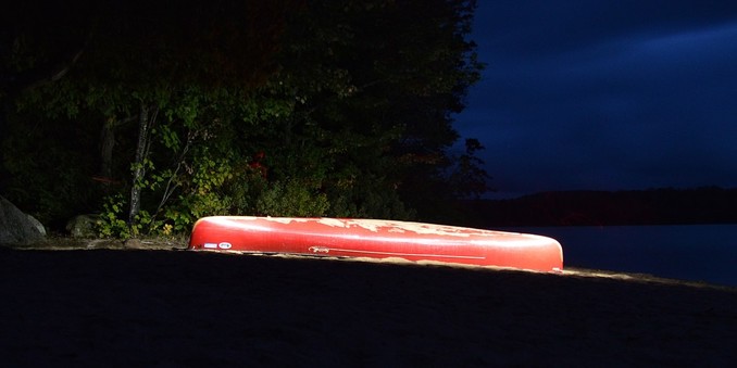 A photo of a red canoe upside-down on a beach. It is night and the canoe has been illuminated by light painting.