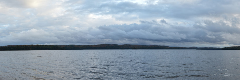 A panoramic photo of a lake. The shores are forest covered hills. The sky is cloudy.