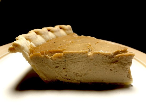 A slice of the aforementioned pawpaw pie on a white plate with beige trim sitting in front of a black background. The slice is in profile and is pointing to the right. This is so you can see what the insides look like. The pie has an orangey hue on top with a dark beige center. The lighting is bright so the slice casts a dark shadow on the white plate. It is a vegan version of cheesecake so it has a similar consistency. Very smooth, custardy but holds form. 