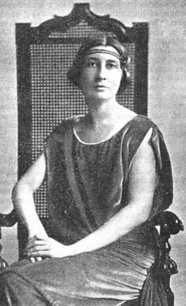 Lilias Mackinnon, from a 1923 publication; a white woman seated in a chair, wearing a headband across her forehead, and a loose velvet tunic-style dress
