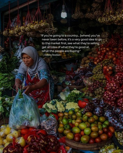 A closeup of a woman working in a market stall, surrounded by vegetables. She is handing a large bag of produce to someone out of frame. The quote on the photo reads, "I’ve also said it over and over again: if you’re going to a country, particularly in Southeast Asia, [where] you’ve never been before, it’s a very good idea to go to the market first, see what they’re selling, get an idea of what they’re good at, what the people are buying." by Anthony Bourdain.