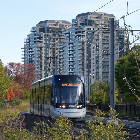 A photo of a light rail train moving on tracks with some trees at their sides. There are two apartments in the background.