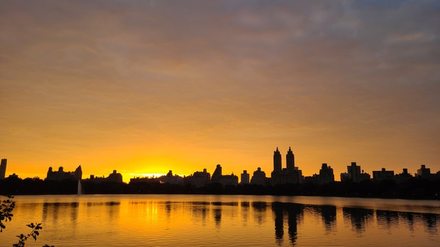 Sunset on the Jacqueline Kennedy Onassis reservoir. The sky is orange and the water of the reservoir is also orange. The silhouette of the Central Park West buildings divides the sky from the water 