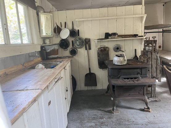 An old cabin interior showing sink and counter, wood cooking stove, some pots and pans on the wall, bunk beds in the back right, white painted walls, and hardwood floor. 
