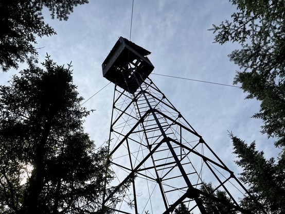 View from bottom of tall metal fire tower. Cables descend  apparently keeping it upright. As view is mostly straight up from below, background is slightly cloudy blue sky encircled by trees. 