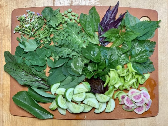 On a wooden cutting board is an assortment of leaves, peas, seed pods, cucumbers, radish, peppers ￼and flowers. 