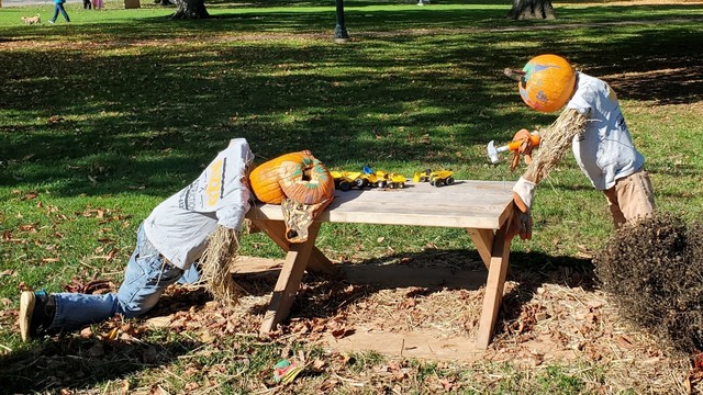 Pumpkin man holding hammer at a table in the park. Other Halloween man lies at other end with pumpkin head smashed in.