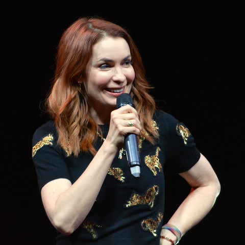 A photo of Felicia Day wearing a black t-shirt with sparkly gold cats on it and holding a microphone. The background is black.