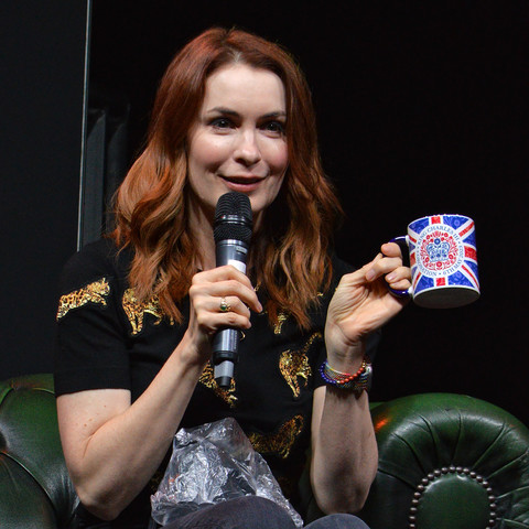A photo of Felicia Day holding up a mug with a union jack pattern on it that says "King Charles III - Coronation - 6th May 2023". She is wearing a black t-shirt with sparkly gold cats on it and holding a microphone.