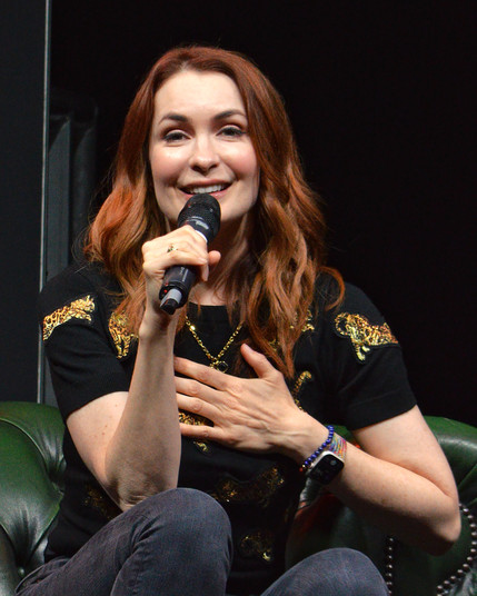A photo of Felicia Day with her hand to her chest. She is wearing a black t-shirt with sparkly gold cats on it and holding a microphone. She is sitting on a green couch.