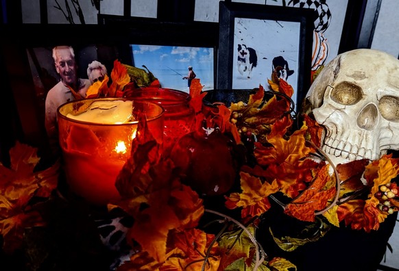 A small table covered in a black cloth holds two lit candles in jars, a small cast iron cauldron, a ceramic pomegranate, and a replica human skull. A garland of fall leaves is arranged between them. Along the back are three pictures in black frames: my maternal grandparents posing together, my father standing on the beach looking at the ocean and holding a fishing rod, and my two dogs running next to each other on snow.