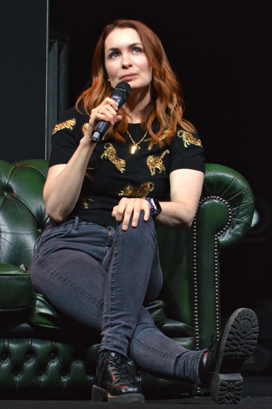 A photo of Felicia Day wearing a black t-shirt with sparkly gold cats on it and holding a microphone. She is sitting on a green couch.