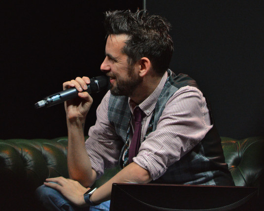 A photo of CJ Allan holding a microphone and sitting on a green couch.
