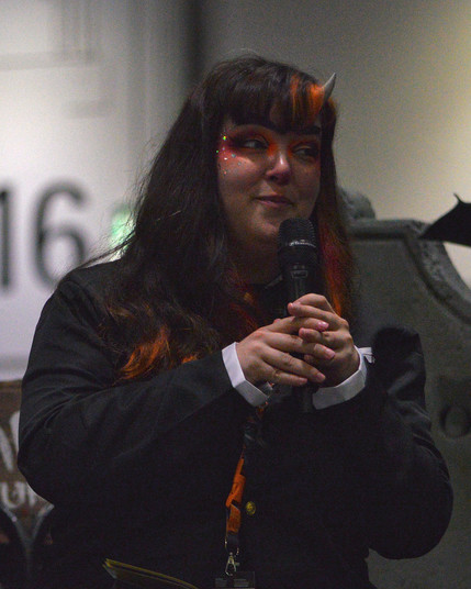 A photo of Taylor Garcia van Biljon holding a microphone. She has orange makeup and sparkles around her eyes, and thick makeup eyebrows. She has a short horn coming out of her forehead.
