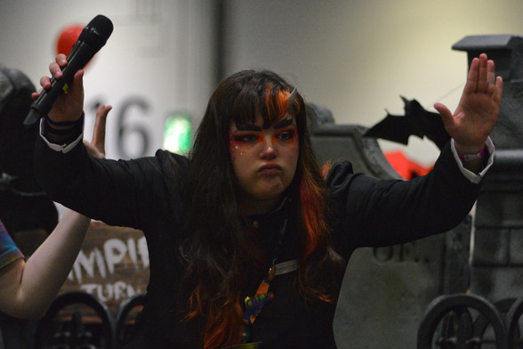 A photo of Taylor Garcia van Biljon with her arms raised in the air one of which is holding a microphone. She has orange makeup and sparkles around her eyes, and thick makeup eyebrows. She has a short horn coming out of her forehead.