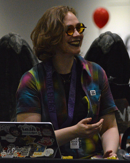 A photo of Fiona K. T. Howat wearing a tie-dye patterned shirt, yellow tinted glasses, and dark lipstick.