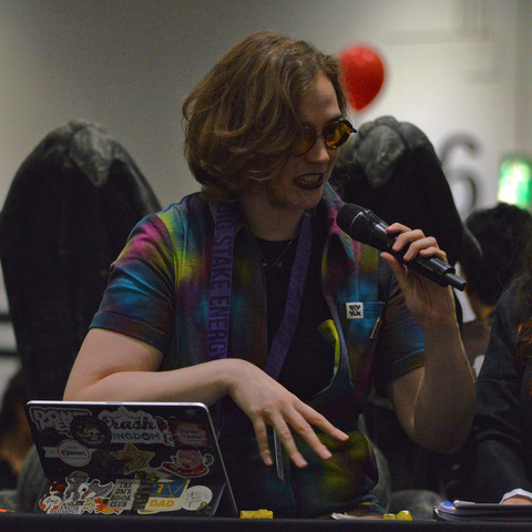 A photo of Fiona K. T. Howat holding a microphone and wearing a tie-dye patterned shirt, yellow tinted glasses, and dark lipstick.