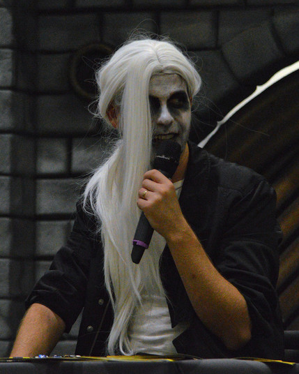 A photo of Hamilton (The Dragon DM) wearing a long white wig and white face paint with dark eyes and holding a microphone.