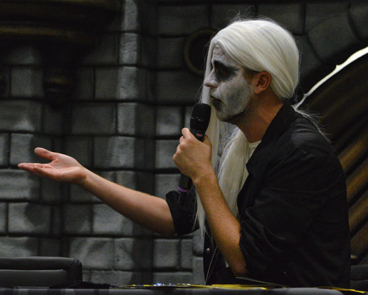 A photo of Hamilton (The Dragon DM) wearing a long white wig and white face paint with dark eyes and holding a microphone with his other hand extended.