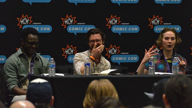 A photo of Drakoniques, Edward Spence, and Fiona K.T. Howat sitting a table covered in a black tablecloth. The board behind them has the logo for "MCM Comic Con" repeating on black.