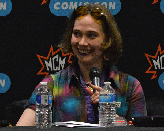 A photo of Fiona K.T. Howat wearing a tie-dye patterned collared shirt. She has yellow tinted glasses pushed up on her forehead. She has dark lipstick on and is wearing large citrus slice earrings. She is holding a microphone and smiling.
