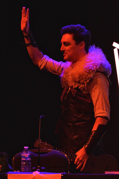 A photo of Luke Westaway as Dob the Half-Orc Bard. He is standing and holding his lute in one hand and waving to the crowd with the other.