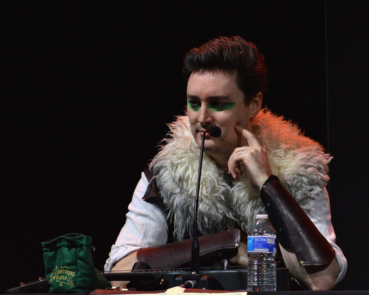 A photo of Luke Westaway as Dob the Half-Orc Bard. He has one finger resting on his cheek as he looks toward the crowd.