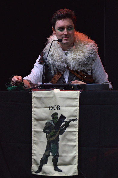 A photo of Luke Westaway as Dob the Half-Orc Bard. He is sitting at a table covered in a black tablecloth. The table has a cloth with his character's drawing and name on it in front of him.