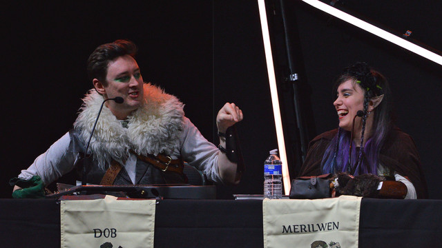 A photo of Luke Westaway as Dob the Half-Orc Bard and Ellen Rose as Merilwen the Wood-Elf Druid. Dob is gesturing with one of his fingers and Merilwen is looking at Dob and laughing.