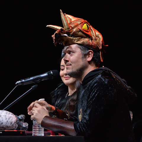 A photo of Mike Channell as Egbert the Careless the Dragonborn Paladin. He has a microphone in front of him and a crazed expression on his face.