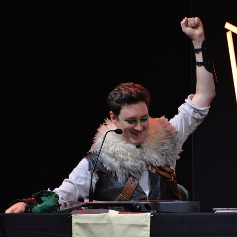 A photo of Luke Westaway as Dob the Half-Orc Bard. He has one arm raised high in the air with his hand closed in a fist. 