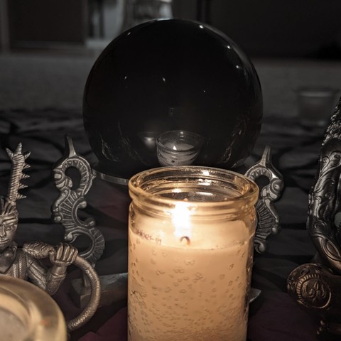 A candle in a jar reflects on an onyx sphere in a stand. To either side, parts of statues of the horned god Cernunnos and a depiction of Mother Earth are within the frame. The image colors have been faded to give a dark feel.