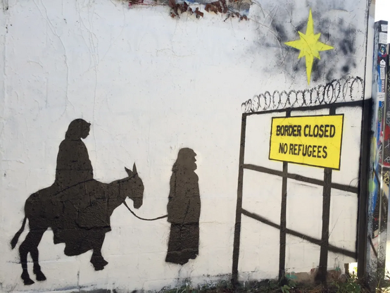 Wall Graffiti:
Painted silhouettes of Joseph guiding Maria sitting on a donkey, stopped by a fence with barbed wire which has a panel "BORDER CLOSED NO REFUGEES". In the sky a comet is shown. 